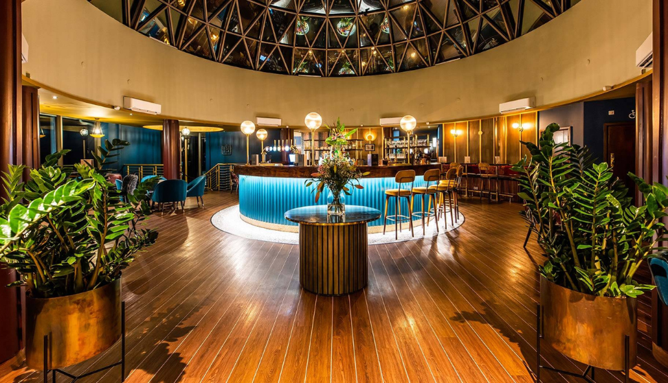 Interior of a stylish circular bar with wooden floor, blue seating, warm lighting, and large plants beneath a glass dome ceiling.